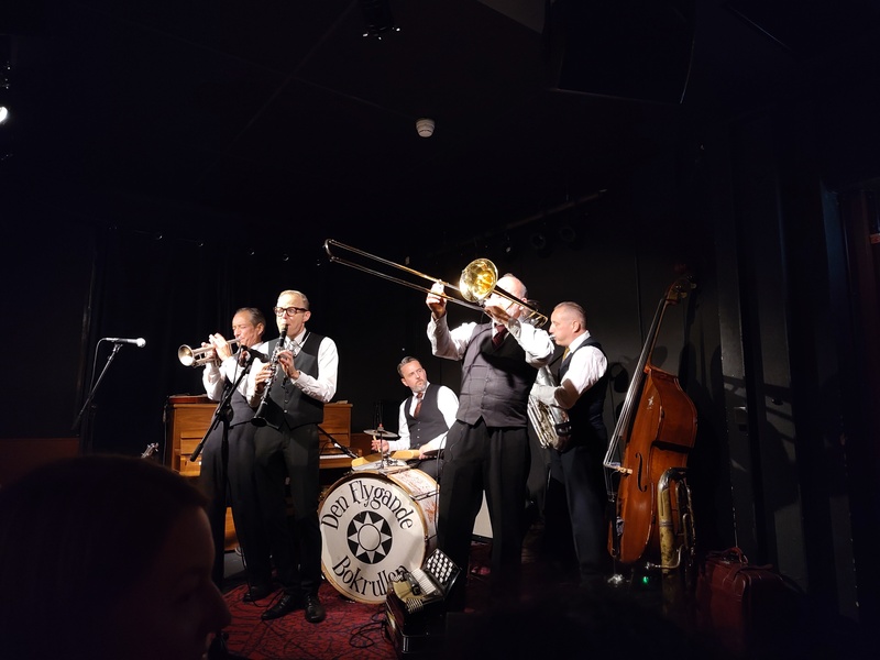 Flere finkledde menn med forskjellige instrument står sammen på en scene og spiller musikk. På trommeskinnet på basstromma står det 'Den flygande bokrullen'.