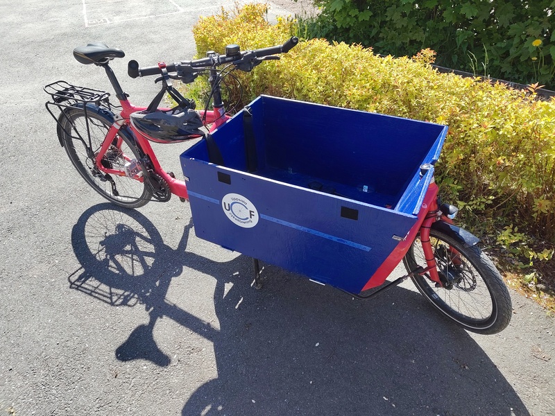 Rød lastesykkel med en blåmalt trekasse fram. Sykkelen står parkert. Den har to hjul: ett fram og ett bak. Frakt meg hvor du vil lille lastesykkel